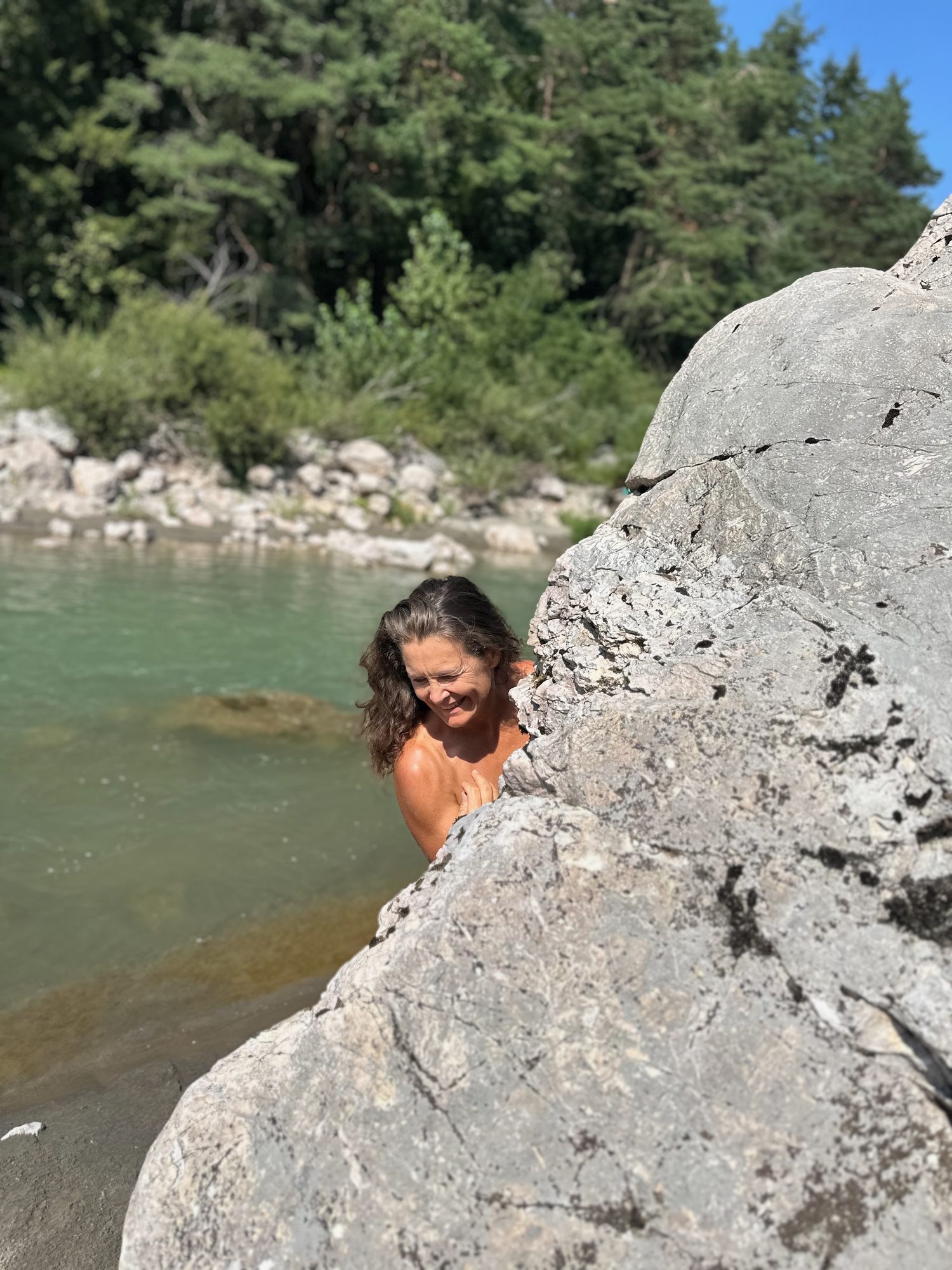 Christina schaut lächelnd hinter einem Felsen am Fluss hervor