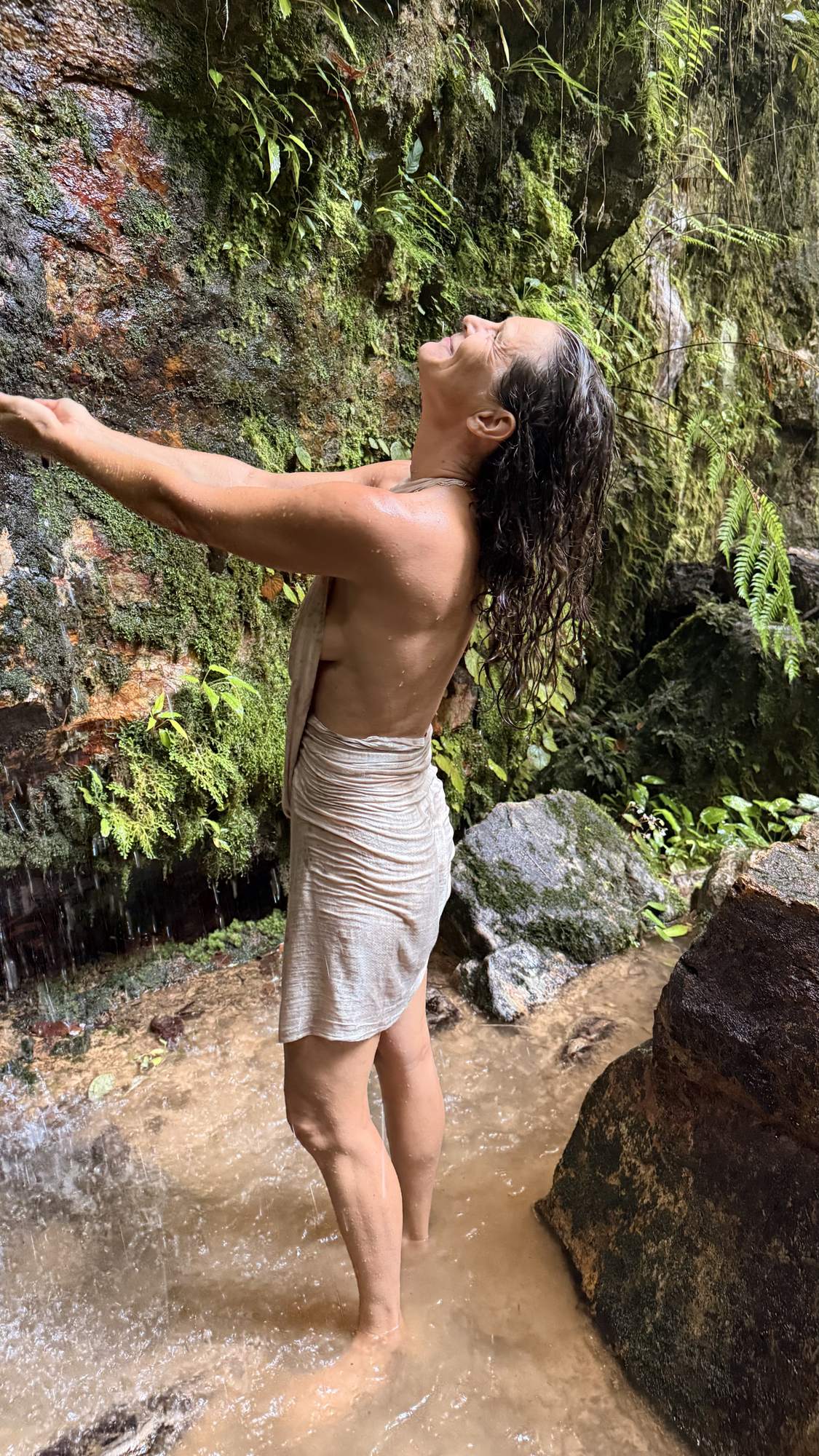 Christina von hinten unter einem Wasserfall, die Arme erhoben